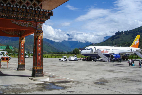 Airplane taxiing on the runway at Paro International Airport in Bhutan, with a second aircraft parked nearby, surrounded by forested mountains and a traditional Bhutanese village.