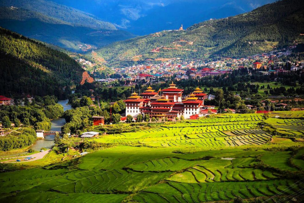 Tashichho Dzong with its whitewashed walls, golden roofs, manicured gardens, and the Wang Chhu River against a Himalayan mountain backdrop.